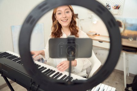 Téléchargez les photos : Smiling joyous young female musician playing synthesizer while looking at smartphone camera - en image libre de droit