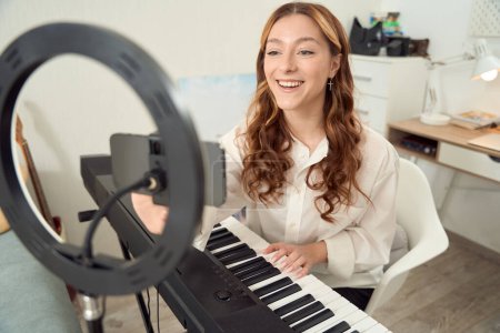 Photo for Smiling pleased young keyboardist sitting at synthesizer and adjusting settings on cellphone - Royalty Free Image