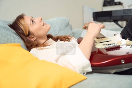 Téléchargez les photos : Smiling happy young female guitarist lying in bed and playing acoustic guitar - en image libre de droit