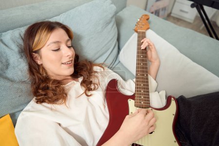 Téléchargez les photos : Smiling pleased young female songwriter involved in playing acoustic guitar in bed - en image libre de droit