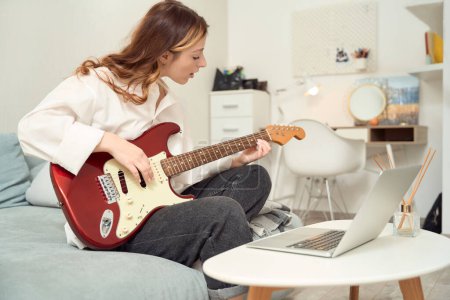Téléchargez les photos : Female seated on bed before laptop brushing fingers over strings of guitar - en image libre de droit