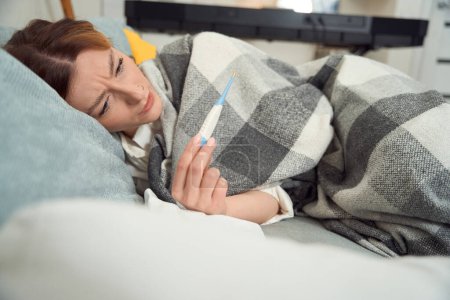 Téléchargez les photos : Worried diseased female wrapped in plaid lying in bed and staring at digital thermometer display - en image libre de droit