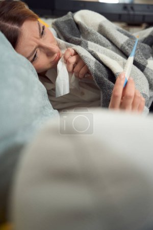 Photo for Sick worried female holding handkerchief and looking at digital thermometer in hand - Royalty Free Image