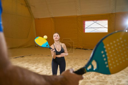 Foto de Focused female in sports uniform practicing hitting tennis ball while playing beach tennis indoors while coach is standing nearby - Imagen libre de derechos
