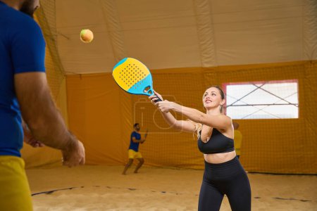 Foto de Encouraged female in sports uniform practicing hitting on sandy beach tennis field while man watching in front - Imagen libre de derechos