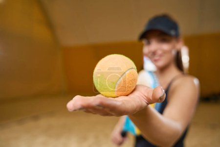 Téléchargez les photos : Smiling female with her hair gathered in a ponytail stands on a sandy field indoors while sports equipment is in her hands - en image libre de droit