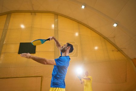 Téléchargez les photos : Focused frescoball player tries to hit the ball with a racket from below while his partner is standing nearby on a sand court - en image libre de droit