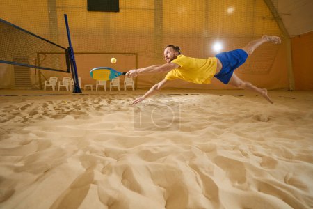 Foto de Enthusiastic tennis player in shorts and t-shirt stretched both arms forward as he falls on the sand - Imagen libre de derechos