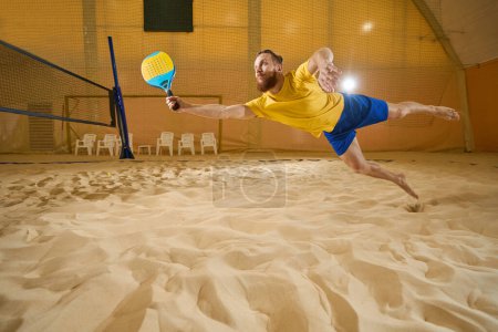Téléchargez les photos : Enthusiastic tennis player in shorts and t-shirt lands on the sand indoors while holding a tennis racket in his hand - en image libre de droit