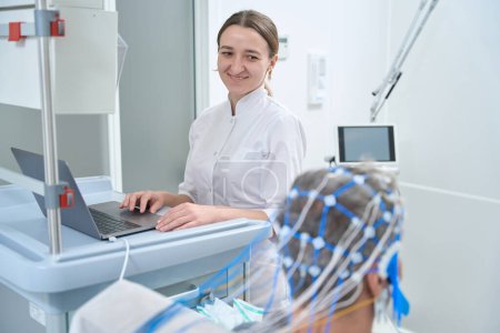 Téléchargez les photos : Friendly female diagnostician performs EEG - electroencephalography, the patient is located in a special chair - en image libre de droit