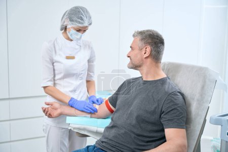 Foto de Smiling patient takes a blood test from a vein, a health worker fixes a patch on a man arm - Imagen libre de derechos