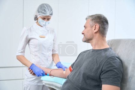 Foto de Health worker taking a blood sample from a smiling man, the patient is sitting in a comfortable medical chair - Imagen libre de derechos