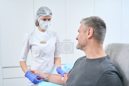 Téléchargez les photos : Nurse in a protective mask takes a blood sample from vein from patient, a man sits in a medical chair - en image libre de droit