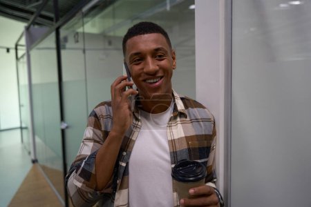 Foto de Happy African American guy talking on smartphone in the office hall and holding coffee - Imagen libre de derechos