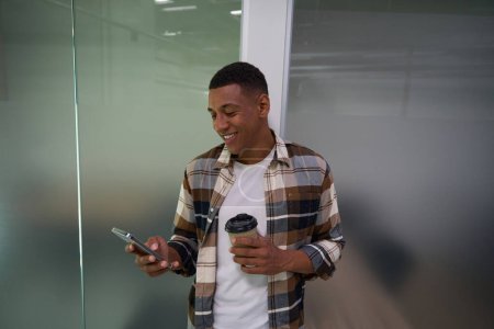 Photo for Happy African American young man typing on mobile phone and holding coffee - Royalty Free Image