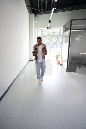 Photo for Full-length of African American young man using mobile phone while walking along office hall, holding cup of drink - Royalty Free Image