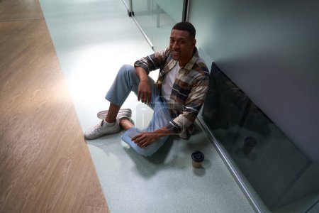 Foto de Top view of happy African American guy sitting on the floor, holding mobile phone in the office - Imagen libre de derechos