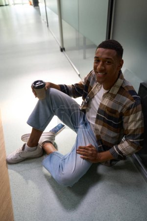 Foto de Smiling African American guy sitting on the floor in the office and holding cup of coffee - Imagen libre de derechos
