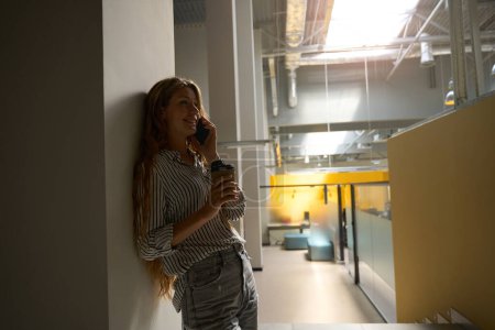 Photo for Happy young lady talking on phone in the office while holding coffee - Royalty Free Image