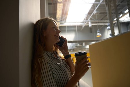 Photo for Side view of smiling young lady talking on mobile phone in the office with cup of coffee - Royalty Free Image