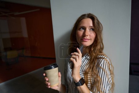Foto de Thoughtful pretty lady holding smartphone and cup of coffee in the office - Imagen libre de derechos