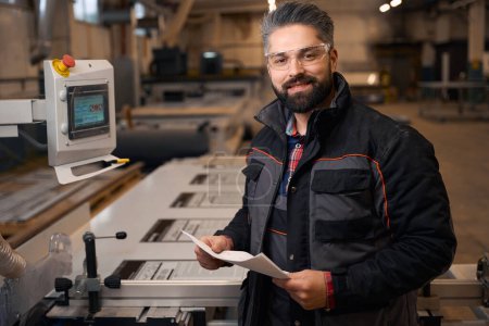 Foto de Smiling man in protective clothes and glasses standing near form cutting machine, holding sheet of paper - Imagen libre de derechos