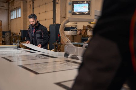 Foto de Man in protective clothes and glasses standing near form cutting machine and cutting material - Imagen libre de derechos