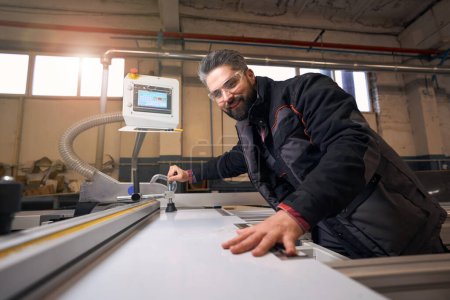 Foto de Portrait of smiling male in protective clothes and glasses standing near woodworking machine and cutting material in workshop - Imagen libre de derechos