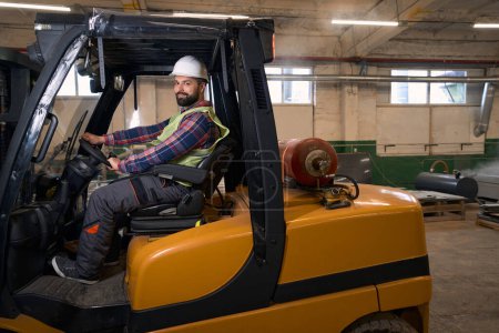 Photo for Smiling man in protective clothes and helmet sitting in the loading machine and driving in manufactory - Royalty Free Image