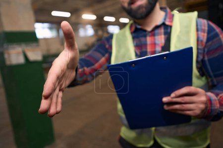 Photo for Male in protective clothes and helmet, standing in workshop, holding clipboard and extend hand to shake - Royalty Free Image