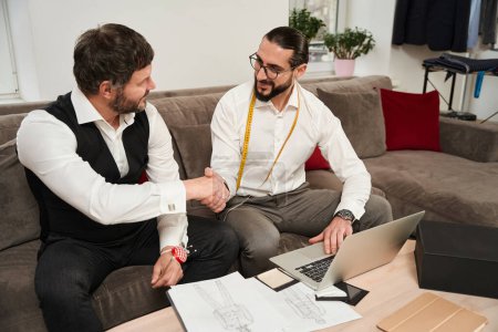 Téléchargez les photos : Pleased couturier sitting at laptop while shaking hands with his business partner - en image libre de droit