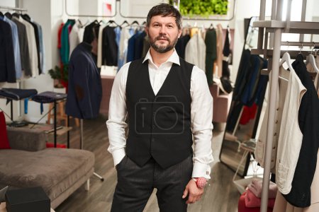 Téléchargez les photos : Calm stylish tailor with wrist-held pincushion posing for camera in his atelier - en image libre de droit