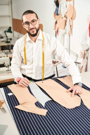 Téléchargez les photos : Tailor with pair of scissors standing at table with pattern pieces arranged on cloth - en image libre de droit