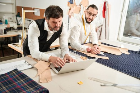 Foto de Clothier arranging pattern pieces on fabric while his colleague typing something on portable computer - Imagen libre de derechos