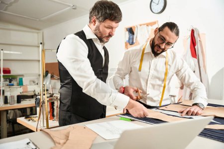 Photo for Serious tailor with chalk and his colleague with pair of scissors standing at cutting table - Royalty Free Image