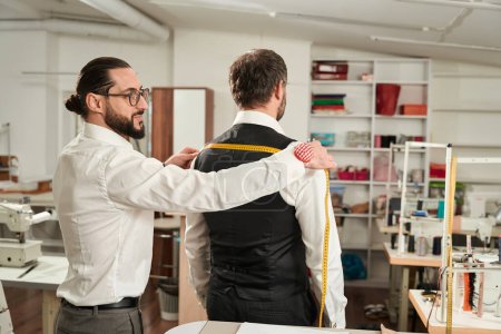 Photo for Focused couturier measuring customer shoulder width using tape measure in his atelier - Royalty Free Image