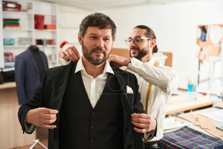 Photo for Adult man putting on custom-made jacket assisted by his pleased personal tailor - Royalty Free Image