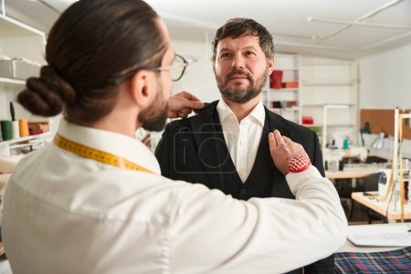 Photo for Experienced couturier checking fit of jacket lapels on adult man in workshop - Royalty Free Image