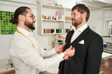 Photo for Clothier testing fit of jacket on his pleased male client in workshop - Royalty Free Image