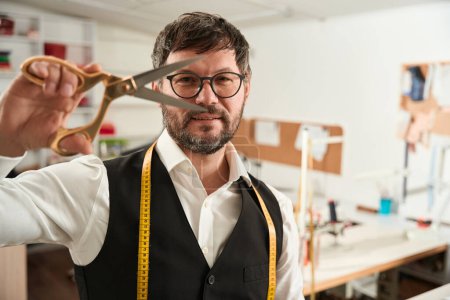 Téléchargez les photos : Waist-up portrait of cheerful couturier with tape measure and pair of scissors in his workplace - en image libre de droit