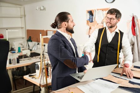 Téléchargez les photos : Smiling pleased fashion designer shaking hands with his colleague at cutting table - en image libre de droit