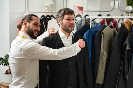 Téléchargez les photos : Smiling contented fashion designer checking fit of jacket collar on his customer - en image libre de droit