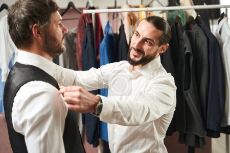 Téléchargez les photos : Smiling contented fashion designer taking male client body measurements in his workshop - en image libre de droit