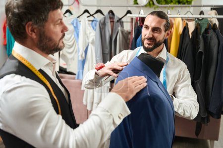 Téléchargez les photos : Joyous clothier and his serious colleague checking fit of jacket on mannequin - en image libre de droit