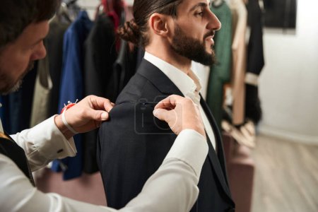 Téléchargez les photos : Serious concentrated tailor pinning jacket sleeve to armhole on his customer during fitting session - en image libre de droit