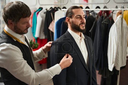 Téléchargez les photos : Calm focused professional tailor adjusting length of jacket sleeve on pleased client - en image libre de droit