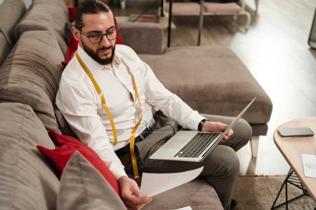 Foto de Serious focused man with laptop and sheet of paper sitting on sofa - Imagen libre de derechos