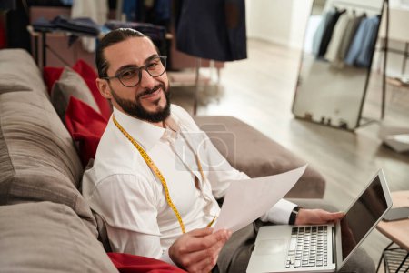 Téléchargez les photos : Smiling male designer sitting on sofa with laptop and clothing sketches - en image libre de droit