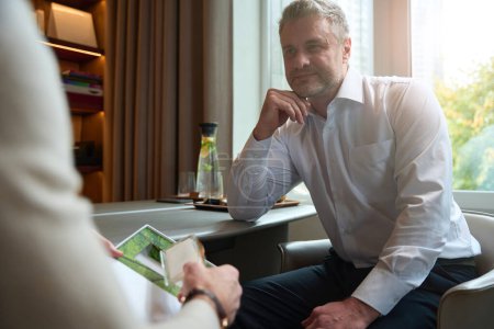 Téléchargez les photos : Waist up portrait of handsome Caucasian man in business clothes is holding hand under the chin while listening his interlocutor in room indoors - en image libre de droit