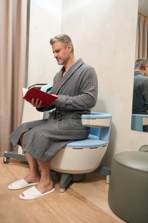 Foto de Retrato de cuerpo entero de hombre barbudo en bata gris está sentado en la silla médica mientras mira a través del libro en la habitación - Imagen libre de derechos